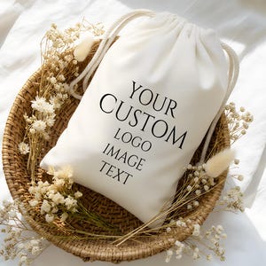 以下が含まれることがあります： 「YOUR CUSTOM LOGO IMAGE TEXT」の黒い文字が入った白い巾着袋。袋は、ドライフラワーで飾られた編みかごの中にあります。背景は白のテクスチャード加工された生地です。