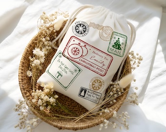 May include: A white drawstring Christmas gift bag with various postage stamp designs in red, green, and gold. The bag includes text that reads "Delivery To: Emily, Christmas Post" and "Do Not Open Until Dec 25th."