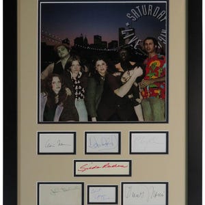 Peut inclure: Photographie encadrée d'un groupe de personnes, avec les mots "SATURDAY NIGHT LIVE 1976" dans un design circulaire. En dessous, plusieurs signatures encadrées. Le cadre est noir.