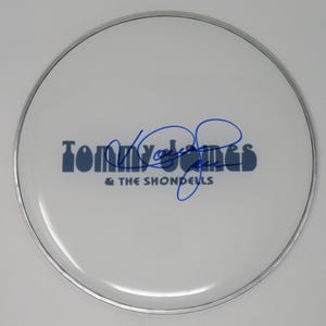 May include: A white drum head with a silver rim. The text "Tommy James & The Shondells" is printed in blue, with a blue signature above the text. The drum head is circular.