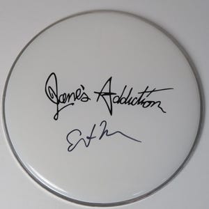 May include: A white drumhead with the band name "Jane's Addiction" and a signature in black ink. The drumhead has a silver-colored metal rim.