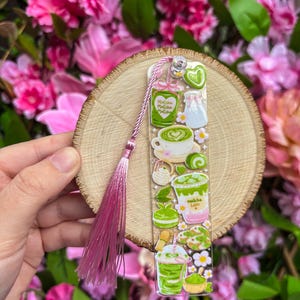 May include: A clear bookmark featuring matcha-themed illustrations, including a cup of matcha latte, a jar, and a matcha drink. The bookmark has a pink tassel and is held against a wooden slice and a backdrop of pink flowers.