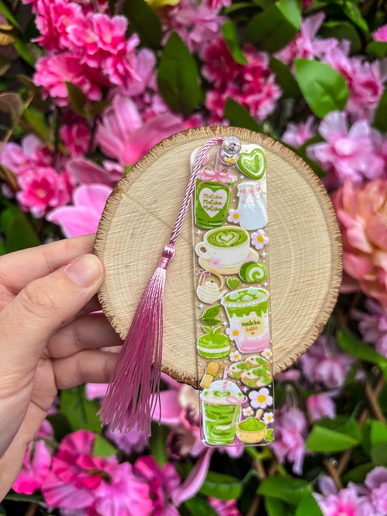 May include: A clear bookmark featuring matcha-themed illustrations, including a cup of matcha latte, a jar, and a matcha drink. The bookmark has a pink tassel and is held against a wooden slice and a backdrop of pink flowers.