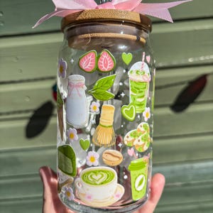 Puede incluir: Un vaso de vidrio transparente con tapa de bamb&uacute; y lazo rosa, decorado con pegatinas tem&aacute;ticas de matcha. Las pegatinas incluyen fresas, bebidas de matcha y una taza de t&eacute;. Una pajita de acero inoxidable est&aacute; insertada en el vaso.