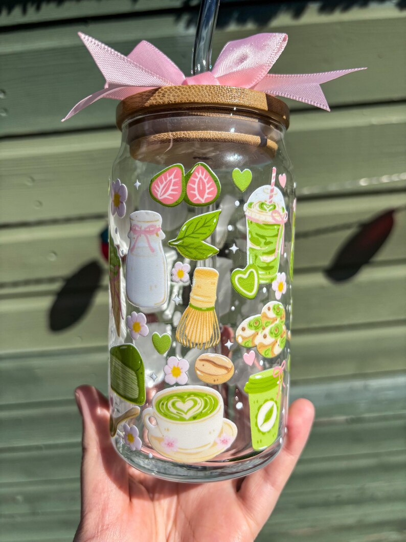 Puede incluir: Un vaso de vidrio transparente con tapa de bamb&uacute; y lazo rosa, decorado con pegatinas tem&aacute;ticas de matcha. Las pegatinas incluyen fresas, bebidas de matcha y una taza de t&eacute;. Una pajita de acero inoxidable est&aacute; insertada en el vaso.