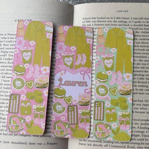 May include: Three decorative bookmarks with a pink and green pattern featuring matcha drinks, pajamas, and floral designs. One bookmark has the name "Lauren" printed on it. The bookmarks are placed on top of an open book.