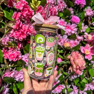 Puede incluir: Un vaso de vidrio transparente con tapa de bamb&uacute;, lazo rosa y pajita, decorado con ilustraciones tem&aacute;ticas de matcha. El vaso se sostiene frente a un fondo de flores rosas. Las ilustraciones incluyen lattes de matcha y otros dise&ntilde;os relacionados con el matcha.
