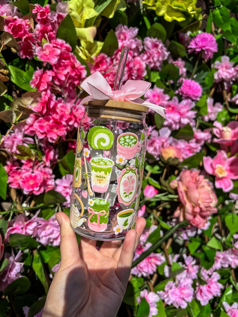 Puede incluir: Un vaso de vidrio transparente con tapa de bamb&uacute;, lazo rosa y pajita, decorado con ilustraciones tem&aacute;ticas de matcha. El vaso se sostiene frente a un fondo de flores rosas. Las ilustraciones incluyen lattes de matcha y otros dise&ntilde;os relacionados con el matcha.