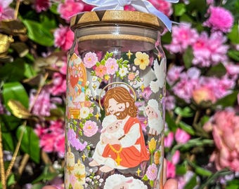 Puede incluir: Un vaso de vidrio con tapa de bambú, pajita transparente y lazo blanco. El vaso presenta una colorida ilustración de Jesús sosteniendo un cordero, rodeado de flores, ovejas y palomas. El fondo es un borrón de flores rosas.
