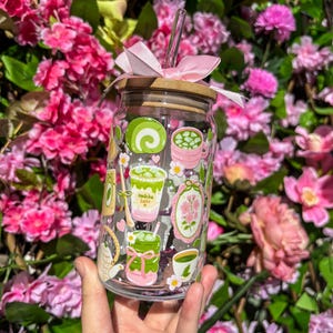 Puede incluir: Un vaso de vidrio transparente con tapa de bamb&uacute; y un lazo rosa. El vaso est&aacute; decorado con ilustraciones tem&aacute;ticas de matcha en verde, rosa y blanco. Una pajita transparente est&aacute; insertada en la tapa. El vaso se sostiene frente a un fondo de flores rosas.