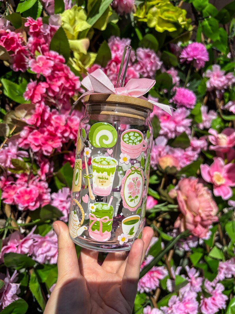 Puede incluir: Un vaso de vidrio transparente con tapa de bamb&uacute; y un lazo rosa. El vaso est&aacute; decorado con ilustraciones tem&aacute;ticas de matcha en verde, rosa y blanco. Una pajita transparente est&aacute; insertada en la tapa. El vaso se sostiene frente a un fondo de flores rosas.