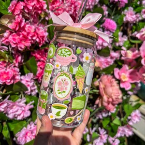 Puede incluir: Un vaso de vidrio transparente con tapa de bamb&uacute; y lazo rosa. El vaso est&aacute; decorado con ilustraciones tem&aacute;ticas de matcha en verde, rosa y blanco, incluyendo lattes de matcha, helado y flores. Se inserta una pajita de vidrio transparente.