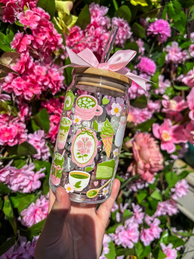 Puede incluir: Un vaso de vidrio transparente con tapa de bamb&uacute; y lazo rosa. El vaso est&aacute; decorado con ilustraciones tem&aacute;ticas de matcha en verde, rosa y blanco, incluyendo lattes de matcha, helado y flores. Se inserta una pajita de vidrio transparente.