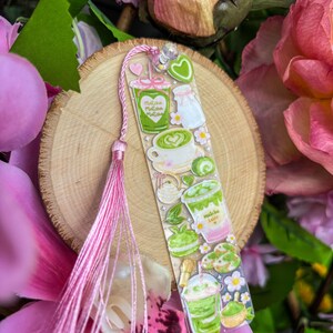 May include: A clear bookmark featuring matcha-themed illustrations, including matcha lattes, macarons, and teacups. The bookmark has a pink tassel and is set against a wooden slice and pink flowers.