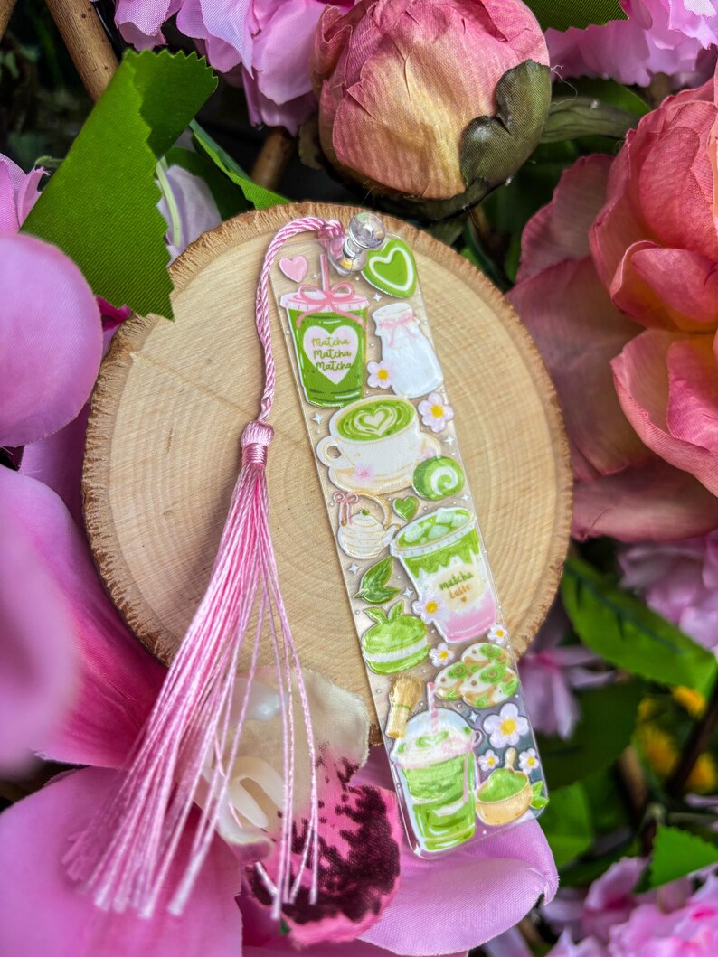 May include: A clear bookmark featuring matcha-themed illustrations, including matcha lattes, macarons, and teacups. The bookmark has a pink tassel and is set against a wooden slice and pink flowers.