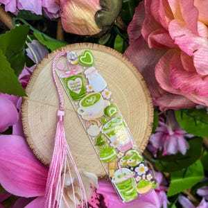 May include: A clear acrylic bookmark featuring matcha-themed illustrations, including drinks, a milk bottle, and flowers. The bookmark has a pink tassel and is set against a wooden slice and pink floral background.