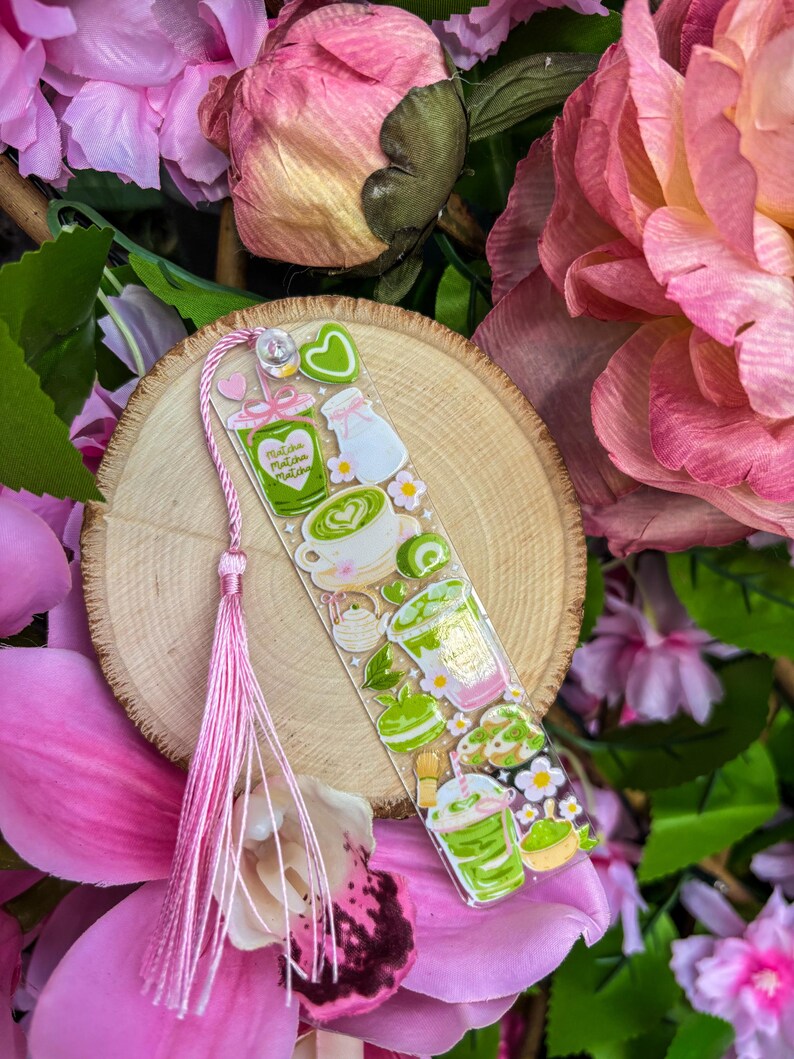 May include: A clear acrylic bookmark featuring matcha-themed illustrations, including drinks, a milk bottle, and flowers. The bookmark has a pink tassel and is set against a wooden slice and pink floral background.