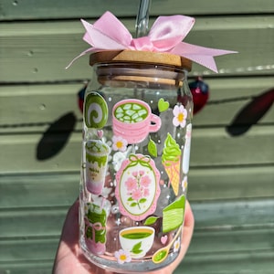 Puede incluir: Un vaso de vidrio transparente con tapa de bamb&uacute; y un lazo rosa. El vaso est&aacute; decorado con pegatinas coloridas con tem&aacute;tica de matcha, que incluyen una taza, helado y pastel. Una pajita de vidrio transparente est&aacute; insertada en la tapa.