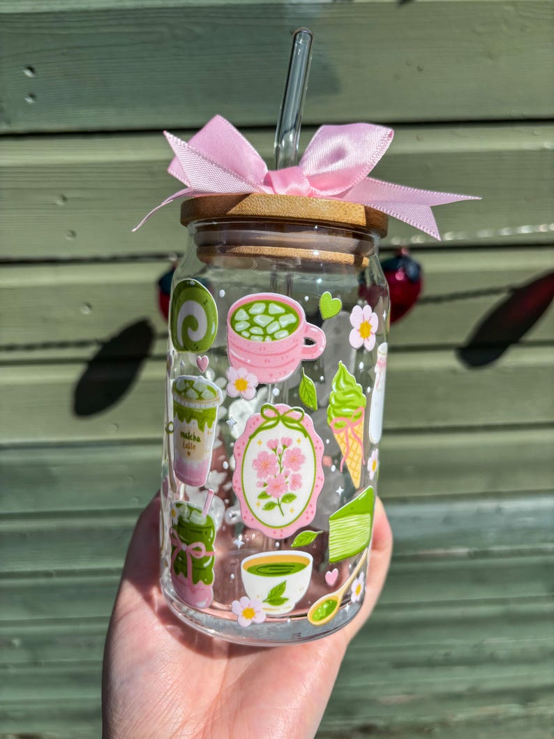 Puede incluir: Un vaso de vidrio transparente con tapa de bamb&uacute; y un lazo rosa. El vaso est&aacute; decorado con pegatinas coloridas con tem&aacute;tica de matcha, que incluyen una taza, helado y pastel. Una pajita de vidrio transparente est&aacute; insertada en la tapa.