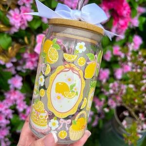 May include: A clear glass tumbler with a bamboo lid, a silver straw, and a white bow. The tumbler is decorated with yellow lemons, white flowers, and a yellow oval frame. The tumbler is designed for cold beverages.