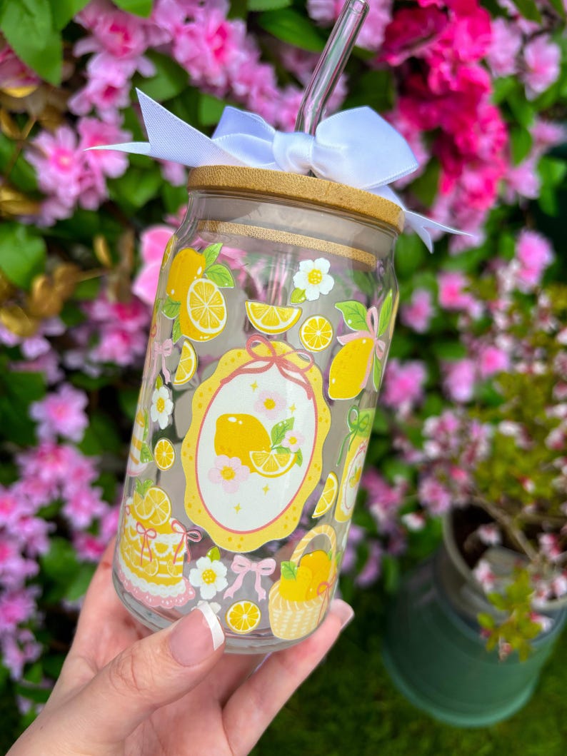 May include: A clear glass tumbler with a bamboo lid, a silver straw, and a white bow. The tumbler is decorated with yellow lemons, white flowers, and a yellow oval frame. The tumbler is designed for cold beverages.