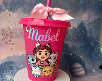 Puede incluir: Un vaso de plástico rosa con tapa y pajita a juego, adornado con un lazo rosa. El vaso presenta un dibujo animado de una niña y gatos, con el nombre "Mabel" en escritura iridiscente.