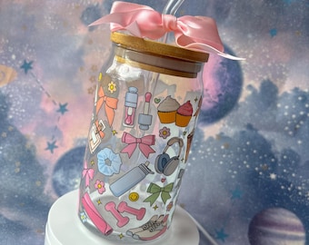 May include: A clear glass tumbler with a wooden lid and a pink bow. The tumbler is decorated with colorful illustrations of bows, makeup, cupcakes, dumbbells, and other accessories. A clear straw is inserted into the tumbler.