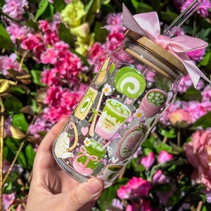 Puede incluir: Un tarro de cristal transparente con tapa de bamb&uacute; y lazo rosa, decorado con pegatinas tem&aacute;ticas de matcha. Las pegatinas incluyen lattes de matcha, un batidor de t&eacute; y un pastel enrollado. Una pajita transparente est&aacute; insertada en el tarro.