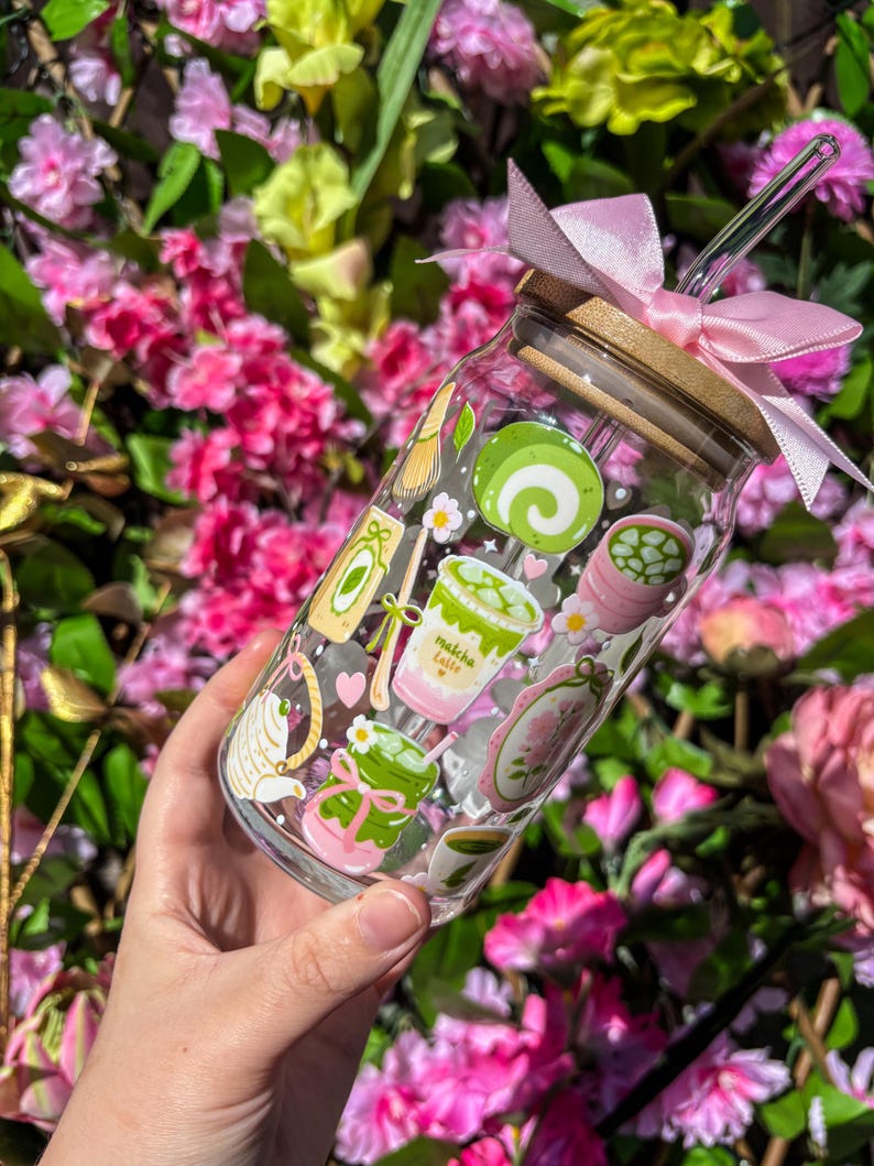 Puede incluir: Un tarro de cristal transparente con tapa de bamb&uacute; y lazo rosa, decorado con pegatinas tem&aacute;ticas de matcha. Las pegatinas incluyen lattes de matcha, un batidor de t&eacute; y un pastel enrollado. Una pajita transparente est&aacute; insertada en el tarro.