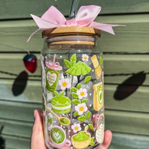Puede incluir: Un vaso de vidrio transparente con tapa de bamb&uacute; y lazo rosa. El vaso est&aacute; decorado con ilustraciones tem&aacute;ticas de matcha, incluyendo un latte de matcha, un batidor y flores. Incluye una pajita de vidrio.