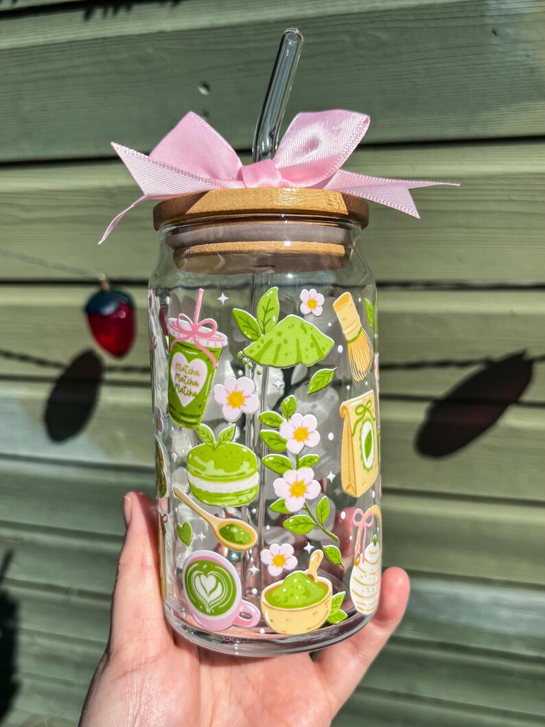 Puede incluir: Un vaso de vidrio transparente con tapa de bamb&uacute; y lazo rosa. El vaso est&aacute; decorado con ilustraciones tem&aacute;ticas de matcha, incluyendo un latte de matcha, un batidor y flores. Incluye una pajita de vidrio.