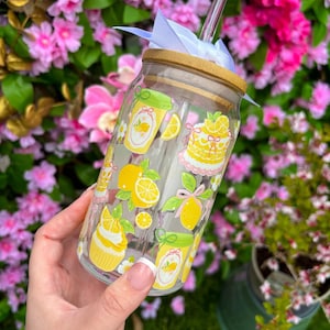 May include: A clear glass tumbler with a bamboo lid and straw, decorated with yellow lemon and dessert illustrations. The tumbler is adorned with a white ribbon and is held against a backdrop of pink flowers.