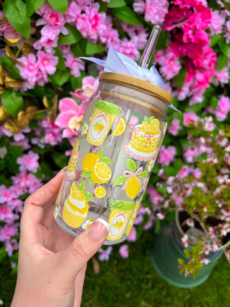 May include: A clear glass tumbler with a bamboo lid and straw, decorated with yellow lemon and dessert illustrations. The tumbler is adorned with a white ribbon and is held against a backdrop of pink flowers.