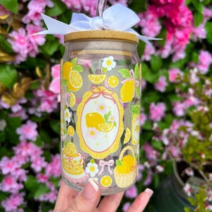 May include: A clear glass tumbler with a bamboo lid, white bow, and glass straw. The tumbler is decorated with yellow lemons, flowers, and a lemon-themed design. The tumbler is approximately 6 inches tall.