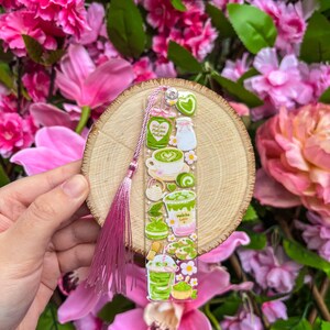 May include: A clear bookmark featuring matcha-themed illustrations, including lattes and desserts, with a pink tassel. The bookmark is held against a wooden slice and surrounded by pink flowers.