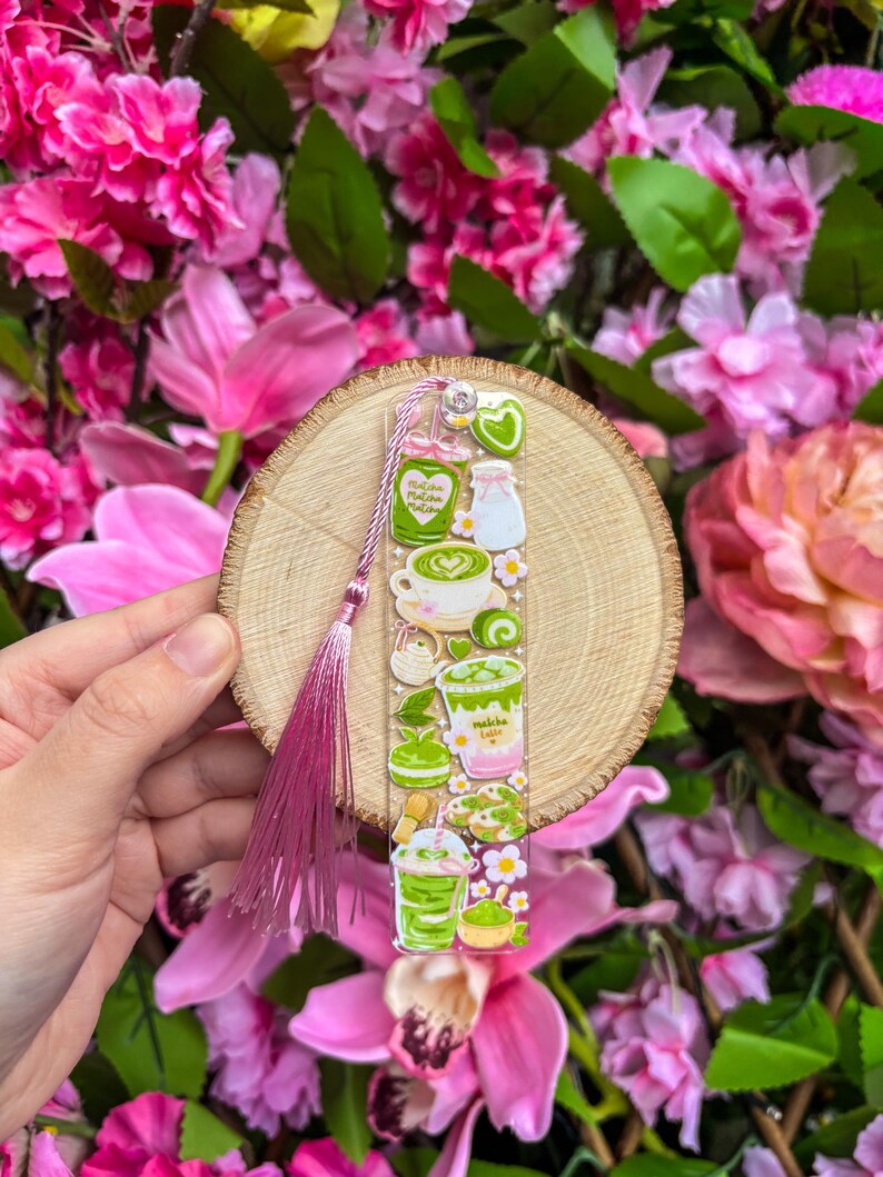 May include: A clear bookmark featuring matcha-themed illustrations, including lattes and desserts, with a pink tassel. The bookmark is held against a wooden slice and surrounded by pink flowers.