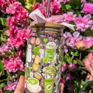 Puede incluir: Un vaso de vidrio transparente con tapa de bamb&uacute; y un lazo rosa. El vaso est&aacute; decorado con pegatinas tem&aacute;ticas de matcha, que incluyen fresas, corazones y bebidas de matcha. Una pajita de vidrio transparente se inserta en el vaso.
