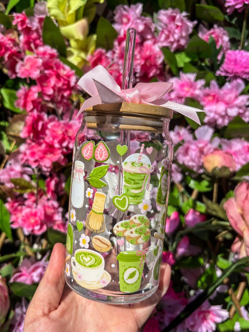 Puede incluir: Un vaso de vidrio transparente con tapa de bamb&uacute; y un lazo rosa. El vaso est&aacute; decorado con pegatinas tem&aacute;ticas de matcha, que incluyen fresas, corazones y bebidas de matcha. Una pajita de vidrio transparente se inserta en el vaso.