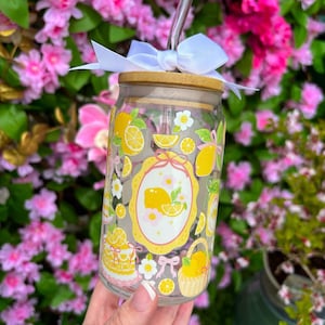 May include: A clear glass tumbler with a bamboo lid and a silver straw. The tumbler is decorated with yellow lemons, white flowers, and a lemon-themed design. A white bow is tied around the lid. The tumbler is held in front of a background of pink flowers.