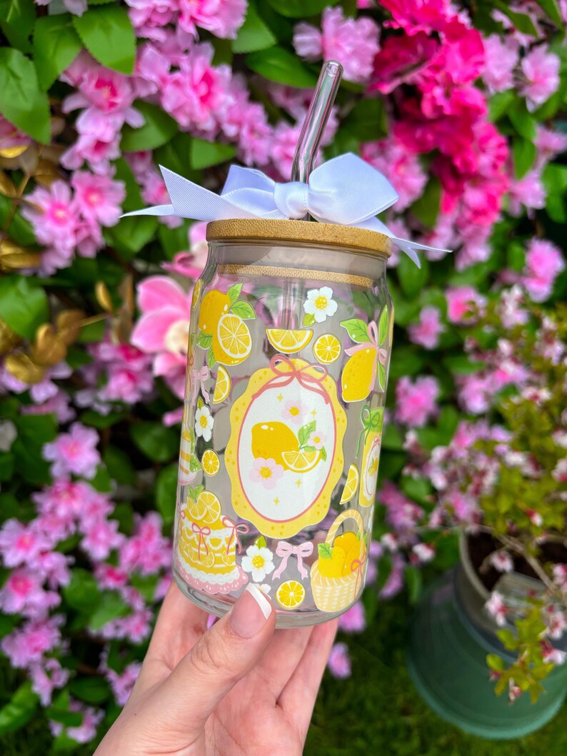 May include: A clear glass tumbler with a bamboo lid and a silver straw. The tumbler is decorated with yellow lemons, white flowers, and a lemon-themed design. A white bow is tied around the lid. The tumbler is held in front of a background of pink flowers.