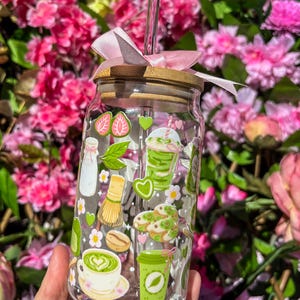 Puede incluir: Un vaso de vidrio transparente con tapa de bamb&uacute; y lazo rosa. El vaso est&aacute; decorado con pegatinas de tem&aacute;tica matcha verde y blanco, incluyendo lattes de matcha, galletas y un batidor. Una pajita transparente est&aacute; insertada en el vaso.