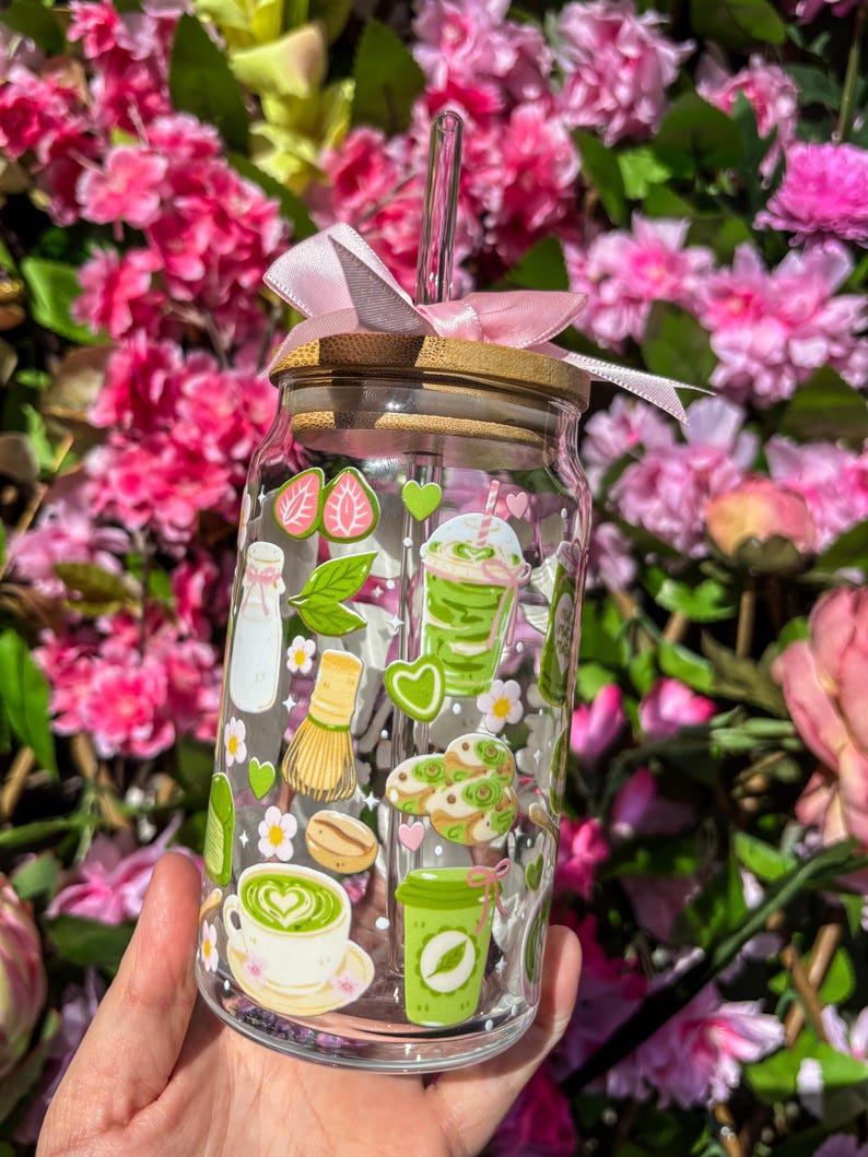 Puede incluir: Un vaso de vidrio transparente con tapa de bamb&uacute; y lazo rosa. El vaso est&aacute; decorado con pegatinas de tem&aacute;tica matcha verde y blanco, incluyendo lattes de matcha, galletas y un batidor. Una pajita transparente est&aacute; insertada en el vaso.