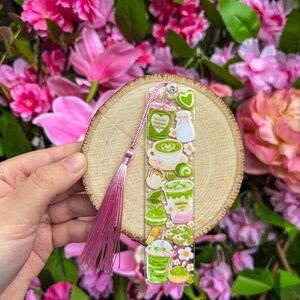May include: A clear acrylic bookmark featuring matcha-themed illustrations, including matcha lattes, cookies, and tea sets. The bookmark is adorned with a pink tassel and a small heart charm. The background is a wooden slice, surrounded by pink flowers.