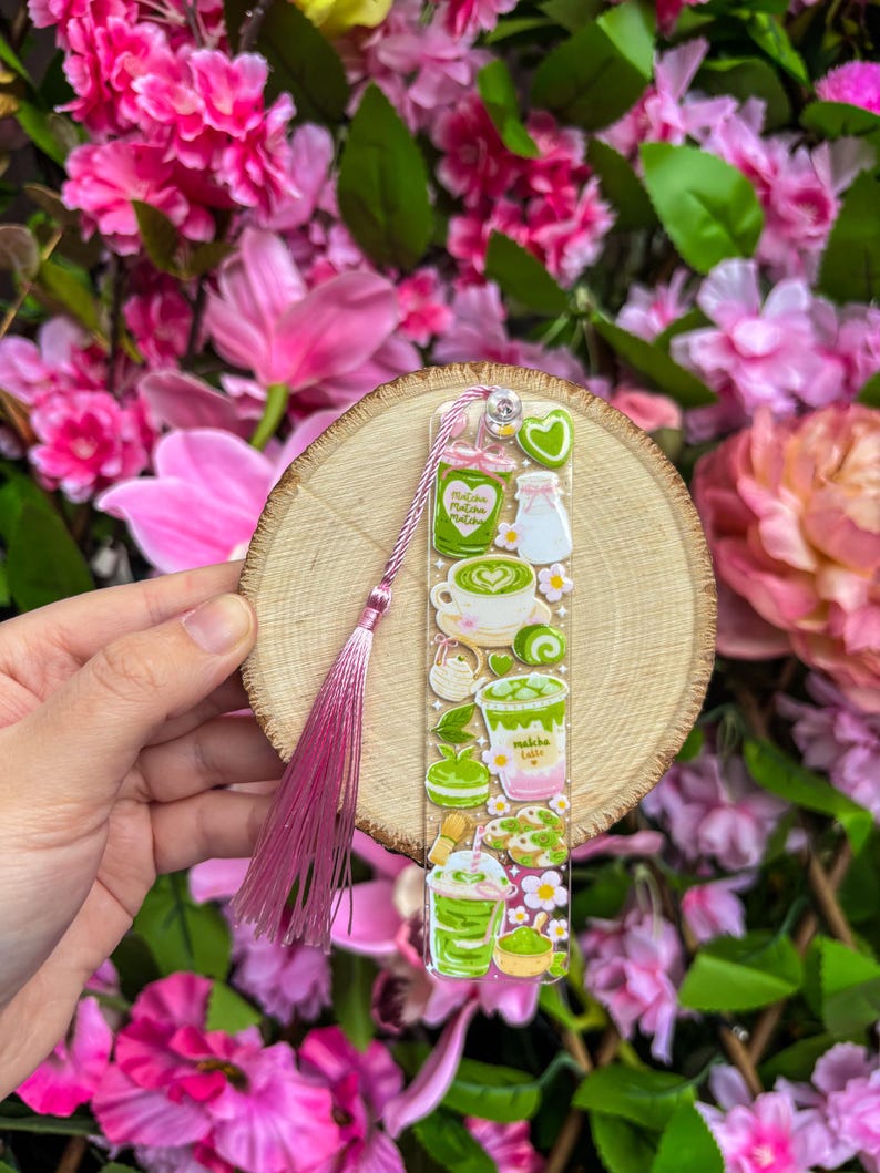 May include: A clear acrylic bookmark featuring matcha-themed illustrations, including matcha lattes, cookies, and tea sets. The bookmark is adorned with a pink tassel and a small heart charm. The background is a wooden slice, surrounded by pink flowers.