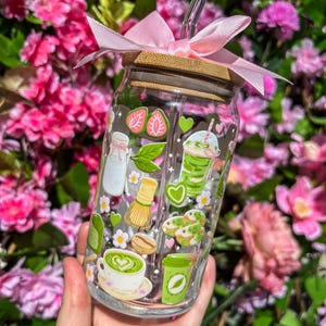 Puede incluir: Un vaso de vidrio transparente con tapa de bamb&uacute; y una cinta rosa atada alrededor. El vaso est&aacute; decorado con pegatinas tem&aacute;ticas de matcha, que incluyen fresas, leche y lattes de matcha. Una pajita de vidrio transparente se inserta en el vaso.