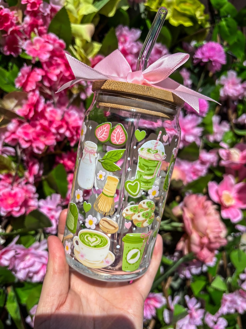 Puede incluir: Un vaso de vidrio transparente con tapa de bamb&uacute; y una cinta rosa atada alrededor. El vaso est&aacute; decorado con pegatinas tem&aacute;ticas de matcha, que incluyen fresas, leche y lattes de matcha. Una pajita de vidrio transparente se inserta en el vaso.