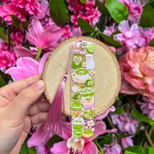 May include: A clear acrylic bookmark featuring matcha-themed illustrations, including lattes, desserts, and ingredients. The bookmark is attached to a wooden disc and a pink tassel. The background is a vibrant display of pink flowers.