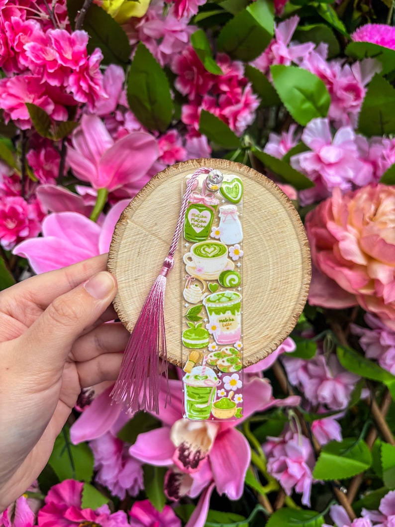 May include: A clear acrylic bookmark featuring matcha-themed illustrations, including lattes, desserts, and ingredients. The bookmark is attached to a wooden disc and a pink tassel. The background is a vibrant display of pink flowers.