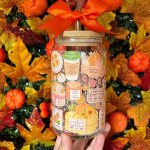 May include: A clear glass tumbler with a bamboo lid and a glass straw, topped with an orange ribbon bow. The glass features colourful autumn-themed illustrations including pumpkins, a coffee cup, a candle, and text like 'Cozy Nights'.