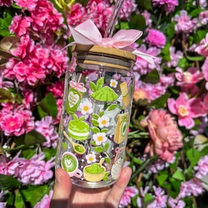 Puede incluir: Un vaso de vidrio transparente con tapa de bamb&uacute; y un lazo rosa. El vaso est&aacute; decorado con ilustraciones tem&aacute;ticas de matcha, que incluyen una taza de matcha, un batidor y un postre de matcha. Una pajita de vidrio transparente se inserta en el vaso.